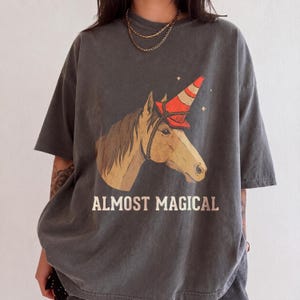 Könnte beinhalten: Ein anthrazitfarbenes Kurzarm-T-Shirt mit einer Grafik eines braunen Pferdes, das einen orange-weißen Verkehrskegel wie ein Horn auf dem Kopf trägt. Unter dem Pferd ist der Text ALMOST MAGICAL in einer cremefarbenen Schrift aufgedruckt.
