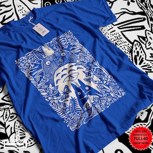 Pu&ograve; includere: T-shirt blu reale con grafica bianca di una scena di foresta con una grande creatura rotonda e figure pi&ugrave; piccole. Il design include farfalle e fogliame. Un'etichetta con la scritta "LARGE" &egrave; attaccata. La maglietta ha scollo tondo e maniche corte.
