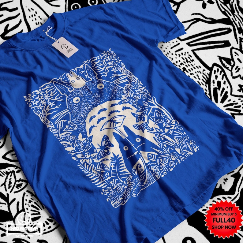Pu&ograve; includere: T-shirt blu reale con grafica bianca di una scena di foresta con una grande creatura rotonda e figure pi&ugrave; piccole. Il design include farfalle e fogliame. Un'etichetta con la scritta "LARGE" &egrave; attaccata. La maglietta ha scollo tondo e maniche corte.