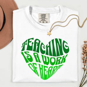 Könnte beinhalten: Ein weißes kurzärmeliges T-Shirt mit einem großen grünen Herzmotiv auf der Brust. Der Text im Inneren des Herzens lautet "TEACHING IS A WORK OF HEART" in einer stilisierten Schriftart. Das T-Shirt hat ein Comfort Colors Etikett am Halsausschnitt.