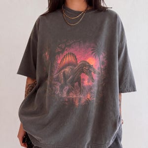 Op de afbeelding: Een antracietgrijs T-shirt met korte mouwen en een grafische print van een Spinosaurus-dinosaurus in een prehistorische jungle. Het kunstwerk toont een levendige oranje zonsondergang, palmbomen en water, met de dinosaurus in het midden.