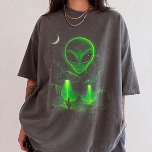 Może przedstawiać: Ciemnoszary t-shirt z krótkim rękawem z wyrazistym zielonym nadrukiem graficznym przedstawiającym dużą głowę kosmity, dwa latające spodki oświetlające pustynny krajobraz z kaktusem oraz sierp księżyca w lewym górnym rogu.
