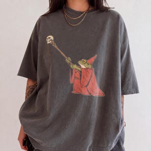 Puede incluir: Una camiseta gris carbón de manga corta con un estampado gráfico de una rana verde con túnica y sombrero puntiagudo rojos, sosteniendo un bastón largo rematado con una pequeña calavera. Tiene cuello redondo y un corte holgado.