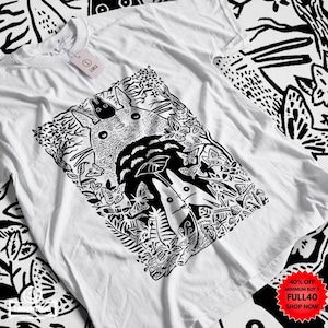 Pu&ograve; includere: T-shirt bianco con grafica in bianco e nero di una scena forestale con una grande creatura rotonda e creature pi&ugrave; piccole. Il design include farfalle e fogliame. La maglietta ha scollo tondo e maniche corte.