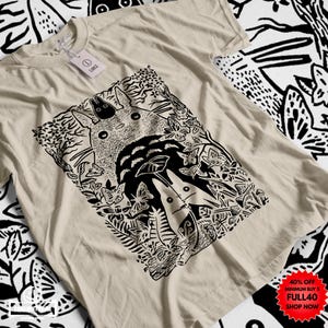 Pu&ograve; includere: T-shirt beige con un'illustrazione in bianco e nero di una scena della foresta, con una grande creatura rotonda e figure pi&ugrave; piccole. Il design include farfalle e fogliame. Una piccola etichetta &egrave; attaccata alla maglietta.