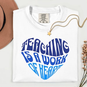 Könnte beinhalten: Ein weißes T-Shirt mit einer blauen herzförmigen Grafik, die den Text "TEACHING IS A WORK OF HEART" in einer wellenförmigen Schrift enthält. Das T-Shirt hat ein Comfort Colors Markenetikett am Kragen, das die Größe XL-XG-TG angibt, 100% Baumwolle.