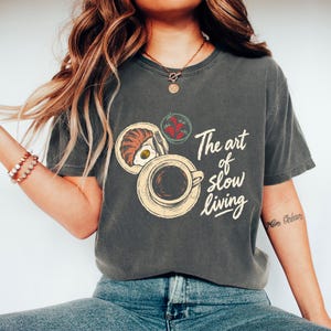 Könnte beinhalten: Ein anthrazitfarbenes T-Shirt mit einem Grafikdruck einer Frühstücksszene mit einem Croissant, einem Spiegelei, einer Schüssel Erdbeeren und einer Kaffeetasse. Der Text "The art of slow living" ist in cremefarbener Schreibschrift aufgedruckt.