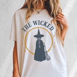 Op de afbeelding: Een wit t-shirt met korte mouwen en een afbeelding van een heks in een donkere mantel en puntmuts die een bezem vasthoudt. Het ontwerp bevat een gele cirkel, kleine sterren en de tekst THE WICKED in een boog boven de centrale figuur.