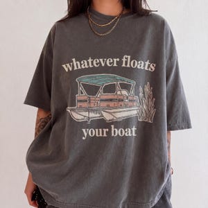 Może przedstawiać: Ciemnoszary t-shirt z krótkim rękawem z grafiką łodzi pontonowej z turkusowym zadaszeniem i różowymi akcentami, otoczonej trzcinami. Napis "whatever floats your boat" jest nadrukowany białymi literami powyżej i poniżej grafiki łodzi.