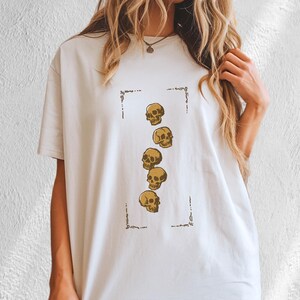 May include: A white short-sleeved t-shirt featuring a central rectangular frame containing a vertical arrangement of five gold-colored human skull illustrations. The graphic is positioned on the chest area of the garment, which has a crew neckline.