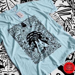 Pu&ograve; includere: T-shirt azzurra con un'illustrazione in bianco e nero di una scena di foresta, con una grande creatura rotonda e figure pi&ugrave; piccole. Il design include farfalle e fogliame. Un'etichetta &egrave; attaccata. La maglietta &egrave; realizzata in materiale morbido.