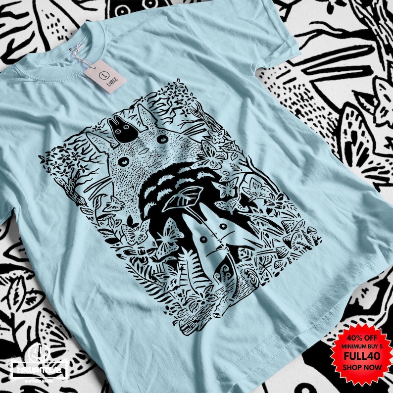 Pu&ograve; includere: T-shirt azzurra con un'illustrazione in bianco e nero di una scena di foresta, con una grande creatura rotonda e figure pi&ugrave; piccole. Il design include farfalle e fogliame. Un'etichetta &egrave; attaccata. La maglietta &egrave; realizzata in materiale morbido.