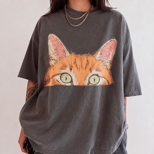 Peut inclure: Un t-shirt gris anthracite à manches courtes orné d'une impression graphique représentant une tête de chat orange aux grands yeux verts regardant au-dessus d'une ligne horizontale. Le t-shirt a un col rond et une coupe décontractée.