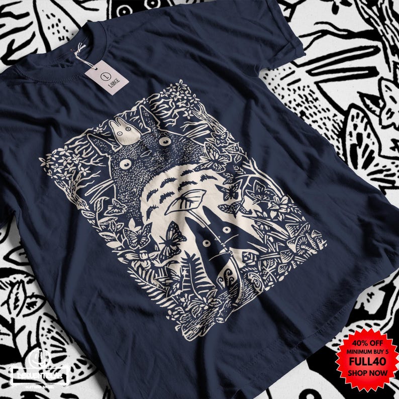 Pu&ograve; includere: T-shirt blu navy con grafica color crema raffigurante una scena di foresta con una grande creatura rotonda e figure pi&ugrave; piccole. Il design include farfalle e fogliame. Un'etichetta &egrave; attaccata. Un adesivo promozionale rosso e bianco &egrave; nell'angolo.