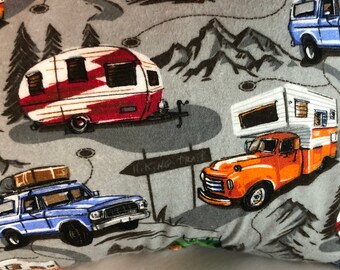 May include: A gray pillow with a camping theme. The pillow features a red, white, and blue camper, a blue truck with luggage on the roof, an orange truck with a white camper, and a green and orange tent. The pillow also has a sign that says "Hiking Trail".