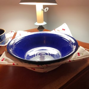 May include: A blue ceramic bowl sits on a white fabric hot pad with a cherry pattern. The hot pad is on a wooden surface. A lamp with a white shade is in the background.