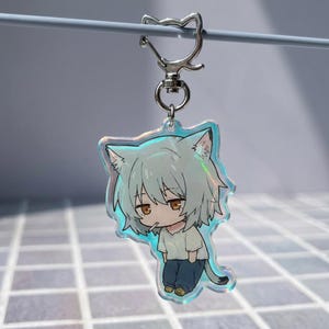 Può includere: Un portachiavi in acrilico raffigurante un personaggio dei cartoni animati con orecchie da gatto, capelli grigio chiaro e occhi ambrati, che indossa una maglietta bianca e pantaloni blu scuro. Il ciondolo è iridescente con una chiusura in metallo.