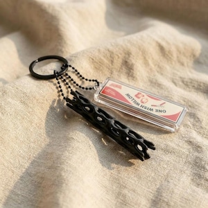 May include: A black keyring featuring a cylindrical, hollow, lattice-patterned charm and a rectangular, clear acrylic pendant. The pendant contains a white insert with red accents and the text ONE WISH WILLOW printed in black lettering.