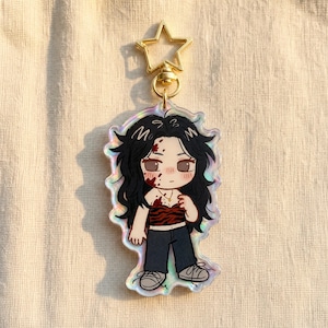 May include: An acrylic keyring featuring a chibi-style character with long black hair, wearing a red and black tiger-striped tube top and dark trousers. The character has red splatters on their face and chest. It includes a gold star-shaped clasp.