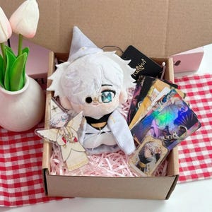 Könnte beinhalten: Eine Plüschpuppe mit weißem Haar und spitzem Hut, bekleidet mit einem weiß-schwarzen Outfit, sitzt in einem Karton. Die Box enthält mehrere rechteckige Karten mit farbigen Illustrationen, einen durchsichtigen Acrylaufsteller und einen Schlüsselanhänger.