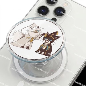May include: A clear phone grip with a white circular design featuring two cartoon cats in robes. One cat is light-coloured, and the other is dark, with gold and blue accents. The grip is attached to a white phone.