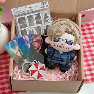 Puede incluir: Una caja de cartón que contiene una muñeca de peluche con cabello rubio y ojos azules, una hoja de pegatinas, tres tarjetas rectangulares con un personaje y un llavero de metal con forma de paraguas rojo y blanco sobre papel rosa.