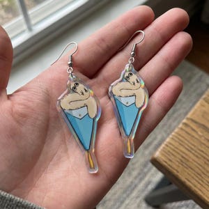 Pode incluir: Um par de brincos pendurados com um desenho de bicho-preguiça de desenho animado. Cada brinco tem uma forma de cone transparente e iridescente com um bolso azul claro e uma preguiça bege. Os brincos têm ganchos de orelha prateados.