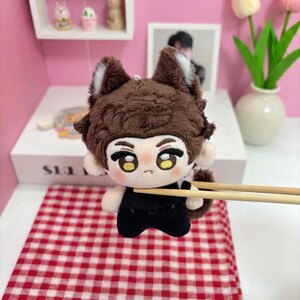 May include: A plush doll featuring brown fur, pointed ears with white inner accents, and large embroidered eyes. The doll has a small mouth and wears a black outfit. It is held by two wooden chopsticks, showcasing its soft texture and detailed facial features.