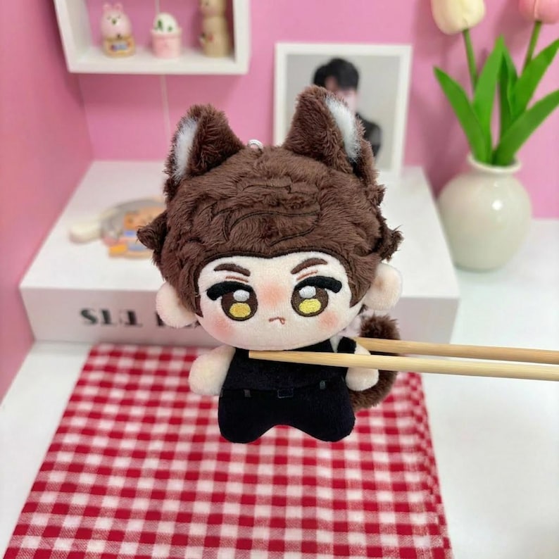 May include: A plush doll featuring brown fur, pointed ears with white inner accents, and large embroidered eyes. The doll has a small mouth and wears a black outfit. It is held by two wooden chopsticks, showcasing its soft texture and detailed facial features.