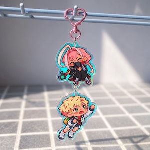 May include: A double-sided acrylic keychain featuring two chibi-style characters. The top charm depicts a character with pink hair and a black mask, while the bottom charm shows a character with blonde hair and a red shirt. Both are connected by a pink heart-shaped clasp.