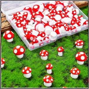 May include: A collection of miniature red and white mushroom figurines with white polka dots. Some mushrooms are scattered on a bed of green artificial grass, while others are inside a clear plastic container. The mushrooms have a glossy finish.