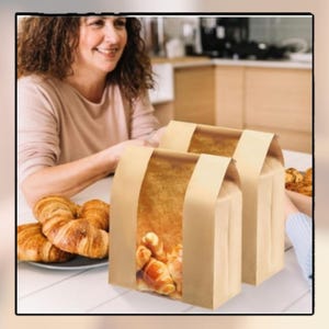 May include: Two brown paper bags with clear windows displaying baked goods, likely croissants, sit on a white table. A plate of golden-brown croissants is in the foreground. A person is visible in the background.
