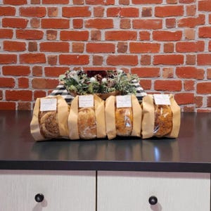 May include: Four loaves of bread in brown paper bags, each with a label, are arranged on a dark countertop. The background features a red brick wall and a decorative arrangement of greenery and pinecones. The bread varieties appear to be different.