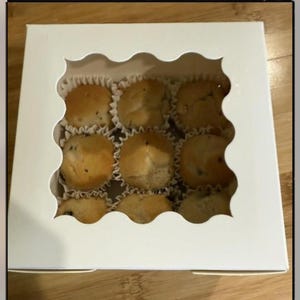 May include: A white bakery box with a scalloped window, showcasing eight mini muffins in paper liners. The muffins are golden brown with dark specks, likely chocolate chips or blueberries. The box sits on a wooden surface.