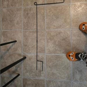 May include: A black metal garden flag holder with a horizontal top bar and a stake for ground insertion. The holder is displayed on a tiled surface. The image also shows a pair of orange and black fox-themed socks.