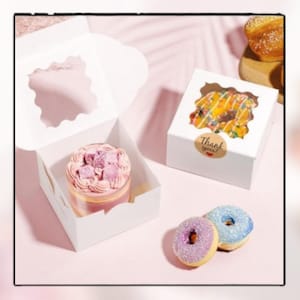 May include: White bakery boxes with clear windows display a frosted cake and a donut. Two donuts, one purple and one blue, sit next to the boxes. A sticker on one box reads "Thank you!"