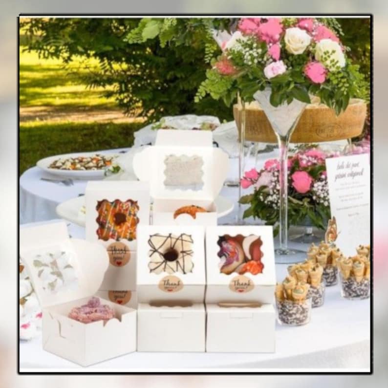 May include: White bakery boxes with clear window tops, each containing a decorated pastry. The boxes are arranged on a white tablecloth, alongside a floral arrangement and small dessert cones. The boxes have "Thank You" stickers.