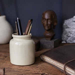 Peut inclure: Un pot à crayons cylindrique en céramique de couleur crème, contenant plusieurs stylos aux finitions noires, brunes et métalliques. Le pot présente un vernis lisse et brillant ainsi qu'un design simple à parois droites avec un bord légèrement évasé.