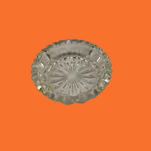 Puede incluir: Un cenicero redondo de cristal transparente con un diseño de estrella en el centro y un borde festoneado. El cenicero es de cristal transparente y se encuentra sobre un fondo naranja.