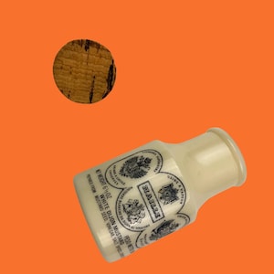 Puede incluir: Una botella de mostaza de Dijon blanca con etiquetas en blanco y negro, y un tapón de corcho, sobre un fondo naranja. La botella tiene la marca "Maille" y el texto "White Dijon Mustard". El peso neto es de 170 gramos.