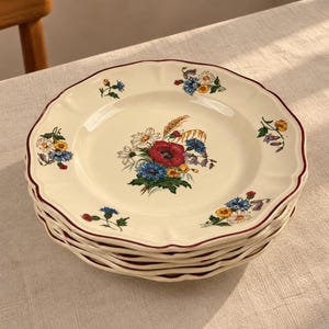 Może przedstawiać: Stos kremowych talerzy ceramicznych z falistymi brzegami i cienką ciemnoczerwoną obwódką. Każdy talerz ozdobiony jest centralnym bukietem kolorowych polnych kwiatów, w tym czerwonych maków, niebieskich chabrów i białych stokrotek.