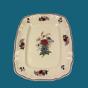 Pode incluir: Prato retangular creme com borda ondulada e acabamento bordô. Apresenta um buquê floral central com papoulas vermelhas, centáureas azuis e trigo. Acentos florais adicionais adornam a borda. Um objeto decorativo.