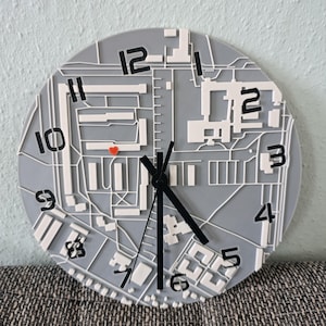 May include: A gray and white wall clock with a map-like design. The clock face features a detailed layout with numbers and black clock hands. A red heart is visible on the clock face. The clock is round and appears to be made of plastic.