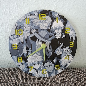 Può includere: Orologio rotondo con un collage di personaggi anime in tonalità di grigio e nero. Il quadrante dell'orologio ha numeri e lancette gialle. L'orologio è su uno sfondo neutro.
