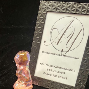 May include: A small, iridescent glass figurine of a kneeling person with hands clasped in prayer. The figurine is in front of a framed sign with the text "AAL Yours Consignments" and a logo.