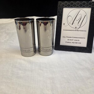 May include: Two cylindrical metallic tumblers with a reflective chrome finish, each featuring the Snap-on logo etched near the base. Beside them stands a black rectangular frame containing a white card with the text AAL Yours Consignments.