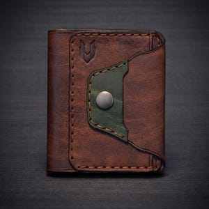 Puede incluir: Una cartera de cuero marrón con un acento verde y un cierre de botón metálico. La cartera presenta un diseño cosido y un pequeño emblema en la parte delantera. El cuero tiene un aspecto texturizado.