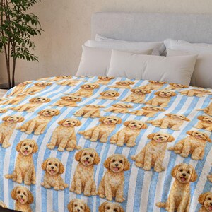 May include: A light blue and white striped blanket featuring a repeating pattern of golden retriever illustrations. The blanket is draped over a bed with white pillows and a gray headboard. The room has a neutral color palette.