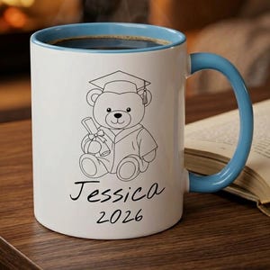 May include: A white ceramic mug with a light blue interior and handle. The front features a black line drawing of a teddy bear wearing a graduation cap and gown while holding a diploma. Below the bear, the name Jessica and the year 2026 are printed.