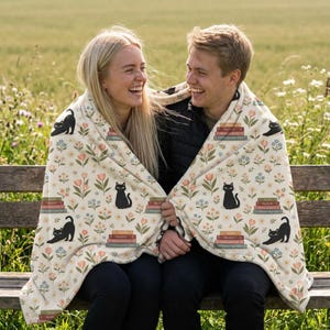 May include: A cream-colored blanket with a floral and cat pattern. The design includes black cat silhouettes, stacks of books, and various flowers in pink, blue, and orange. The blanket is draped over two people sitting on a wooden bench.